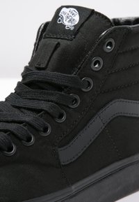 Adidași negri cu guler înalt, din pânză, cu șireturi plate, ochiuri din metal și o talpă din cauciuc texturat de culoare neagră. Au un patch cu logo pe limbă.