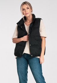 Czarna kamizelka puchowa z wysokim kołnierzem, zatrzaskami i bocznymi kieszeniami, noszona na kremowym swetrze o fakturze, w zestawie z niebieskimi dżinsami.