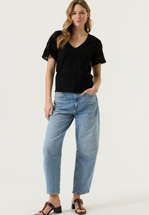 Vrouw draagt een zwart kort mouw kanten top, lichtblauwe broek met hoge taille en bruine sandalen met hakken, staat met één hand in haar zak.