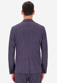 Uomo di spalle che indossa un giacchino scuro a righe verticali viola e pantaloni abbinati su uno sfondo neutro.