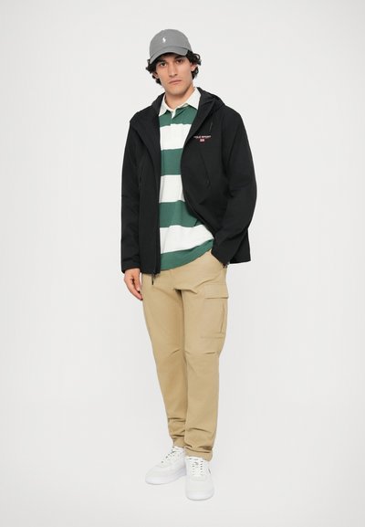 Jachetă neagră cu glugă, tricou polo alb cu dungi verzi, pantaloni cargo bej și adidași albi. Cappă gri cu detaliu de logo.