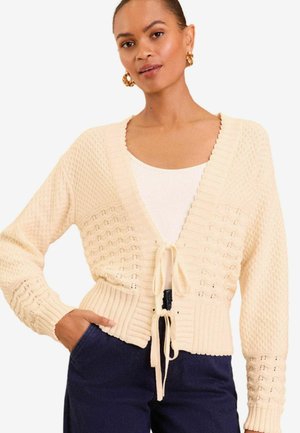 Cardigan tricotat crem, cu un model texturat, decolteu în formă de V și închidere cu fundă. Mânecile și tivul sunt realizate din tricot rib. Purtați peste un top alb și pantaloni albaștri.