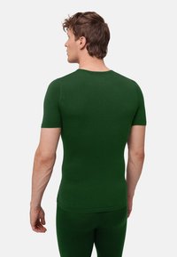 Uomo che indossa una camicia verde scuro aderente a maniche corte e pantaloni coordinati, in piedi di spalle su uno sfondo chiaro e uniforme.