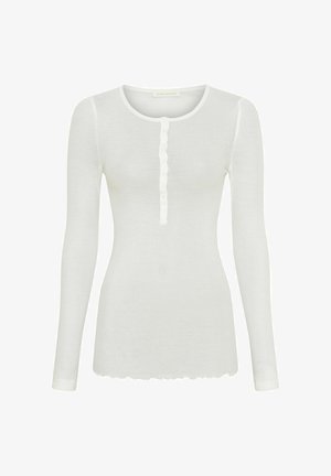 Vit långärmad skjorta i mjukt tyg med rundad halsringning, med en knappslå fram och en scalloped (vågig) nederkant.