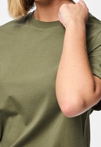 Olivově zelené krátké tričko z bavlněné tkaniny, s kulatým výstřihem a volným střihem, se zřetelným šitím a hladkým povrchem.