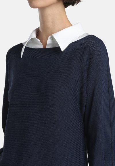 Pulover bleumarin cu design tricotat texturat, având o cămașă albă cu guler dedesubt. Accente nervurate vizibile de-a lungul gâtului și mânecilor.