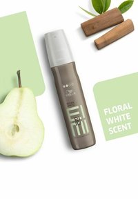 Brun sprayflaska märkt "Wella EIMI Ocean Spritz" bredvid en grön päronskiva och träbitar, på en ljusgrön bakgrund.