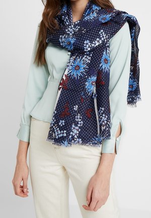 Femme portant un chemisier bleu clair et un pantalon crème avec une écharpe bleu marine ornée de motifs floraux bleus et rouges drapée sur ses épaules.