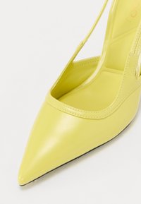 Chaussure à talon haut jaune avec un bout pointu, finition brillante, côtés ouverts et design épuré. Présente des coutures le long des bords.