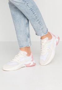 Nogi ubrane w jasnoniebieskie dżinsy z postrzępionymi mankietami oraz białe i różowe sportowe sneakersy na białej podłodze.