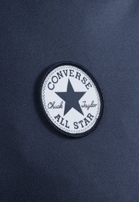 Un patch rond blanc avec une étoile noire et le texte "CONVERSE Chuck Taylor ALL STAR" sur un fond texturé bleu marine.