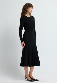 Schwarzes Langarmkleid mit tailliertem Oberteil und ausgestelltendem Rock, aus weichem Stoff gefertigt. Vollendet mit schwarzen Slingback-Schuhen mit spitzem Zeh.