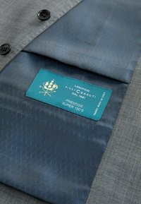 Sivi materijal s glatkom, teksturiranom površinom, s plavim oblogom s dijamantnim uzorkom. Oznaka glasi "Lanificio F.lli Cerruti, Prestige Super 130's."