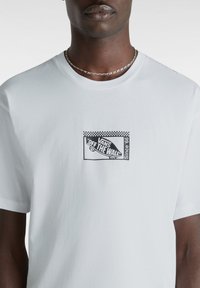 Camiseta de algodón blanca con un estampado gráfico negro que presenta "VANS OFF THE WALL" y una patineta, acentuada por un borde de tablero de ajedrez.