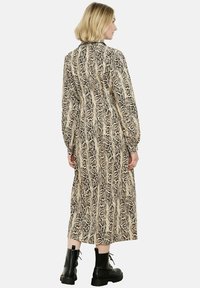 Žena s blond vlasy, oblečená v dlouhých šatech s hadím vzorem a černých šněrovacích kotníkových botách, stojící s otočeným zády.