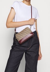 Brązowa torebka w wzory z bordową skórzaną podstawą i paskiem na ramię. Posiada łańcuchowy detal i prostokątny kształt.