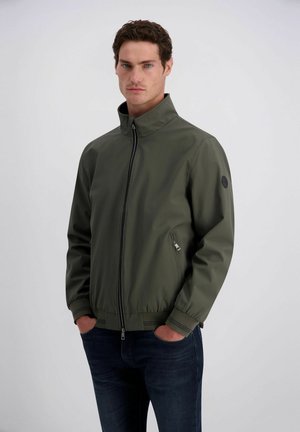 Mann, der eine dunkelgrüne Jacke mit Reißverschluss und hohem Kragen sowie dunkelblaue Jeans trägt, steht mit den Händen in den Taschen vor einem einfarbigen Hintergrund.
