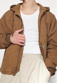 Brun hættejakke med lynlås, frontlomme og snoretræk. Båret over en hvid tanktop, parret med beige shorts med elastisk talje.