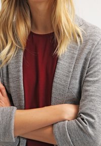 Cardigan grigio con vestibilità comoda, texture a maglia e maniche arrotolate, indossato sopra una maglietta bordeaux. Braccia incrociate davanti.