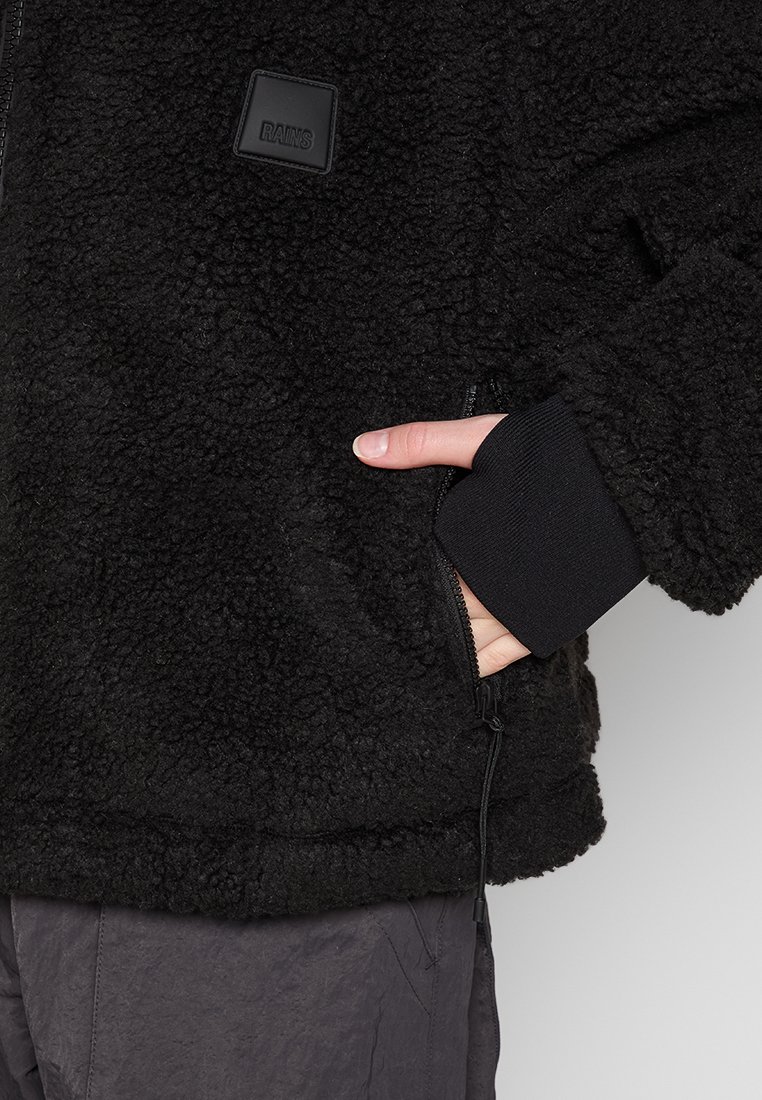 Veste en polaire noire avec une surface texturée et duveteuse, des poignets côtelés et une poche frontale zippée avec une petite étiquette logo.