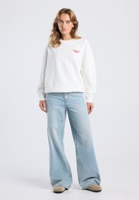 Sudadera blanca con texto bordado rojo, combinada con jeans de pierna ancha en un tono azul claro. Tejido suave con un ajuste relajado y mangas largas.