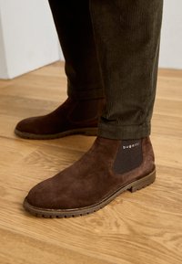 Botas Chelsea de ante marrón con panel lateral elástico y suela de goma, con un acento de marca, llevadas con pantalones de pana oscuros sobre un suelo de madera.