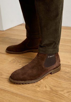Bottines Chelsea en daim marron avec panneaux élastiques et semelle en caoutchouc, arborant un accent de marque, portées avec un pantalon en velours côtelé foncé sur un sol en bois.