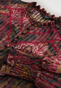 Multicolored blouse with vibrant red, black, and brown patterns; features textured smocking, ruffled neckline, and decorative stitching accents.