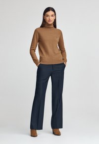 Brązowy sweter z golfem o drobnym splocie z prążkowanymi mankietami, zestawiony z granatowymi spodniami o szerokich nogawkach. Modelka ma na sobie brązowe botki na obcasie.