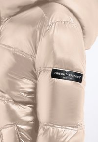 Zbliżenie na błyszczący beżowy rękaw kurtki puchowej z czarną naszą łatą "Frieda & Freddies New York".