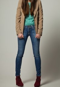 Béžový na míru šitý blazer přes tyrkysový pletený svetr, spárovaný s modrými slim džíny a vínovými vysokými jehlovými kotníkovými botami.