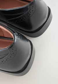 Musta kiillotettu nahkakengät brogue-yksityiskohdilla, matala kanta ja ruskea nahkasisus vaalealla taustalla.