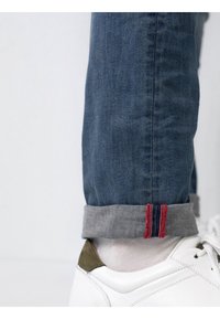 Jeans de mezclilla azul oscuro con dobladillo acampanado que presenta tela gris y acentos de costura en rojo y azul. Combinados con zapatillas blancas.