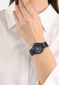 Schwarze Armbanduhr mit einem runden blauen Zifferblatt, goldenen Stundenmarkierungen, einem schwarzen Gehäuse und einem strukturierten Armband, getragen am Handgelenk über einem weißen Hemd.