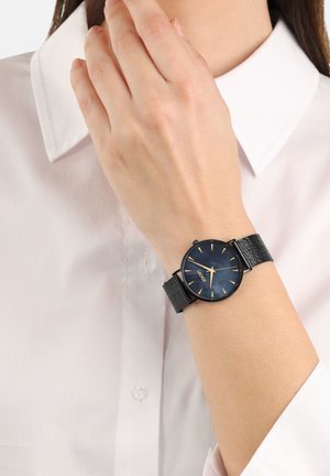 Schwarze Armbanduhr mit einem runden blauen Zifferblatt, goldenen Stundenmarkierungen, einem schwarzen Gehäuse und einem strukturierten Armband, getragen am Handgelenk über einem weißen Hemd.