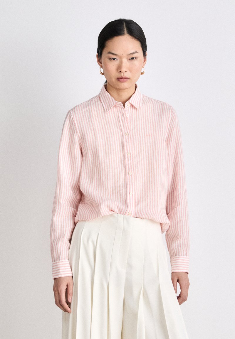 Light pink striped button-up shirt with a collar and long sleeves. Features vertical stripes and subtle textured fabric. Paired with a white pleated skirt.
