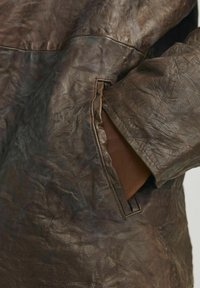 Brown leather jacket with a textured finish, featuring a side pocket and a wide cuff. The material appears slightly crinkled and supple.