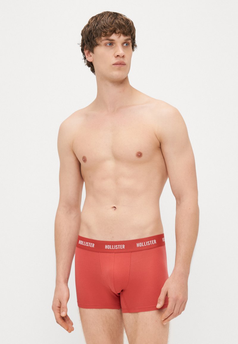 Boxer briefs vermelhos feitos de tecido suave, com uma faixa elástica branca com a marca "Hollister", design ajustado com uma textura lisa.