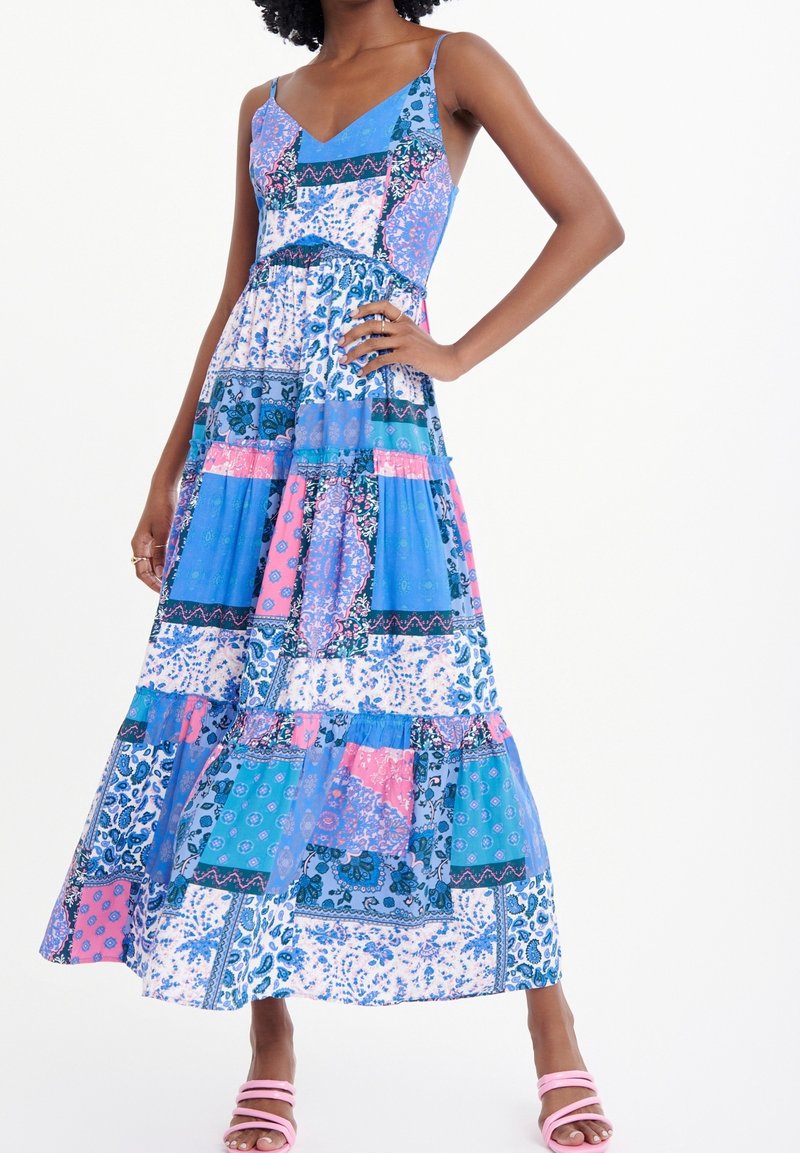 Femme dans une robe maxi sans manches à motifs avec un design en patchwork bleu, rose et blanc, portant des sandales à lanières roses, posant avec la main sur la hanche.