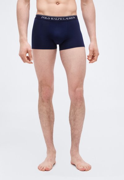 Άνδρας που φοράει σκούρο μπλε ανδρικά εσώρουχα Polo Ralph Lauren, όρθιος ξυπόλητος πάνω σε απλό λευκό φόντο.
