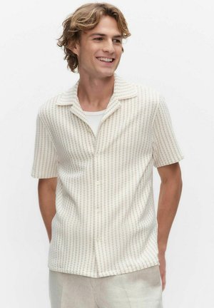 Młody mężczyzna uśmiecha się, nosi teksturowaną, krótką koszulę zapinaną na guziki na białej koszulce i jasnych spodniach.