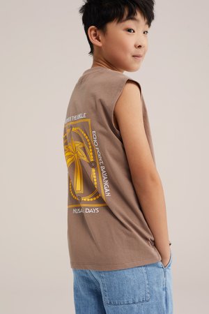 Garçon portant un débardeur marron avec un motif de palmier jaune et un jean bleu clair, regardant sur le côté avec les mains dans les poches.