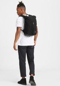 Mochila negra con diseño de rollo en la parte superior, correas ajustables y bolsillos laterales, fabricada con un tejido duradero y acabado texturizado.