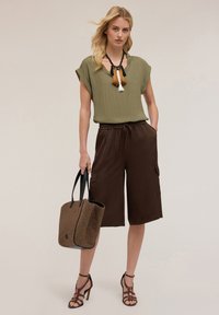 Top verde oliva a maniche corte, pantaloni cargo marroni con vita elasticizzata, borsa intrecciata marrone e nera, sandali alti marroni con cinturini.