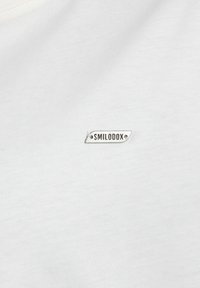 Biała bawełniana koszulka z małą, prostokątną srebrną metką z wypukłym napisem "SMILODOX". Gładka faktura oraz dopasowany krój.