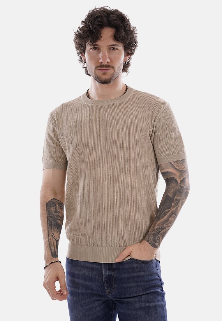 Uomo con capelli ricci che indossa una maglietta a maniche corte beige in maglia e jeans blu, mostra tatuaggi su entrambe le braccia, in piedi contro uno sfondo bianco.