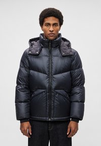 Casaco acolchoado em azul-marinho escuro, com gola alta, fecho de correr e dois bolsos frontais com costuras diagonais e textura suave e brilhante.