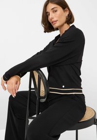 Zwarte lange mouwen top met een ronde halslijn, voorzien van een beige gestreepte accent bij de taille en manchetten. Draagt deze boven een zwarte broek, zittend op een stoel.