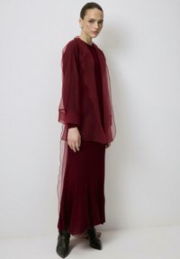 Vestido fluido en tono burdeos con sobreposición translúcida. Presenta una capa base ajustada y tela texturizada, acentuada con mangas anchas y bordes festoneados.