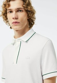 Vit polo skjorta med gröna kontrastsömmar, korta ärmar och en knappslå med tre knappar; har en diskret vit logotyp på bröstet.